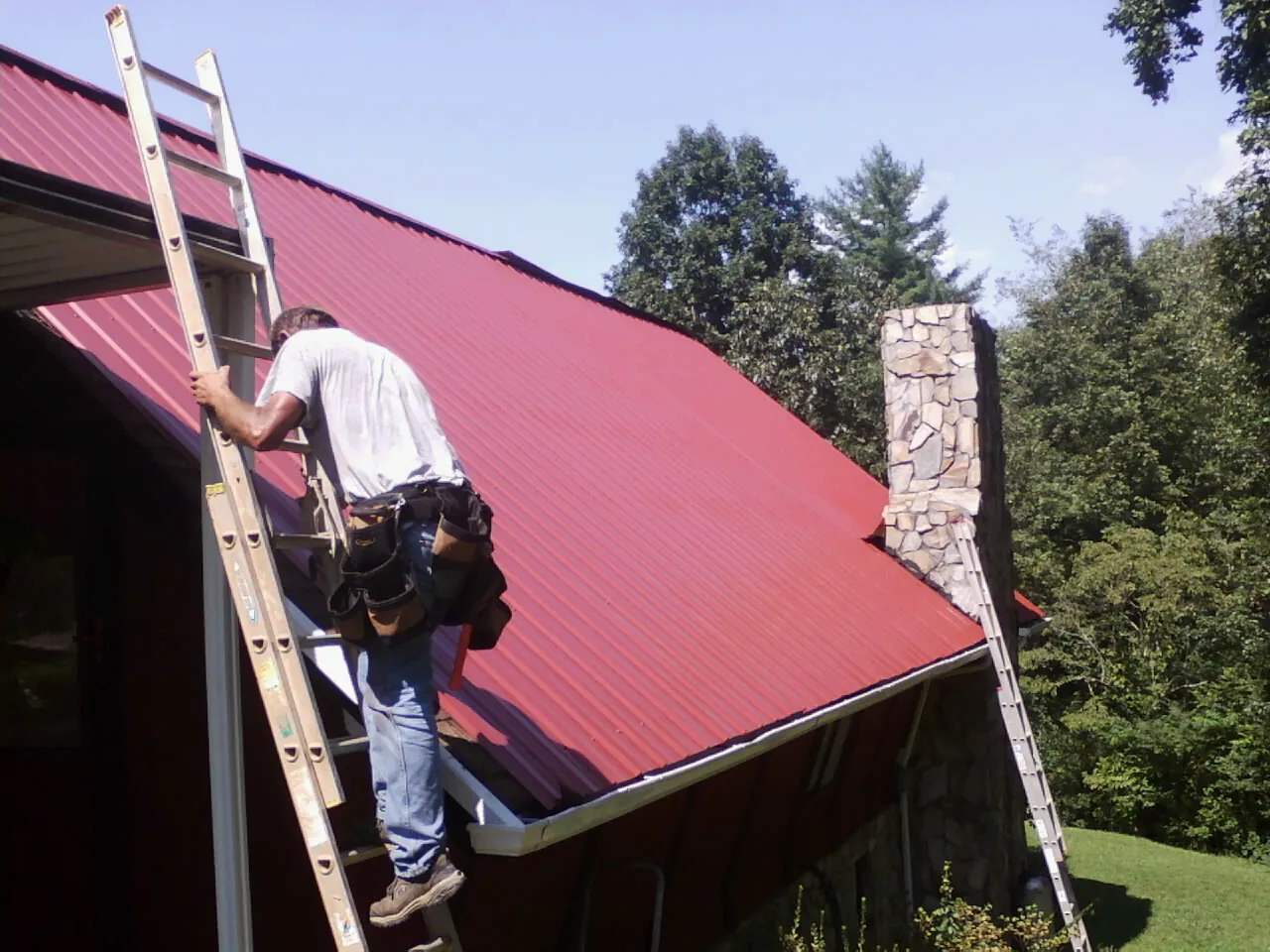 Roofer on a ladder beside a red metal roof and stone chimney — Soffit & Fascia Repair by Roofing Brothers in Scottsville